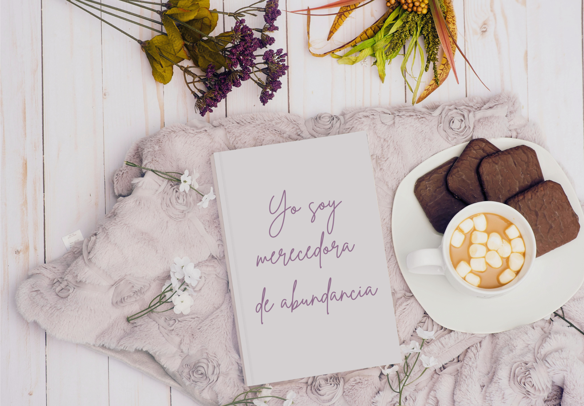 Cuaderno blanco con la afirmación "Yo soy merecedora de abundancia", rodeado de flores, una manta suave, una taza con bebida caliente y galletas de chocolate.